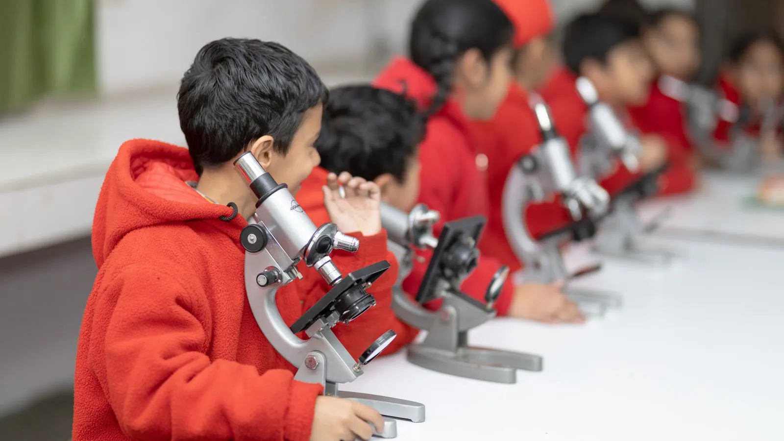 Young students in red jackets looking through microscopes in the junior science lab
