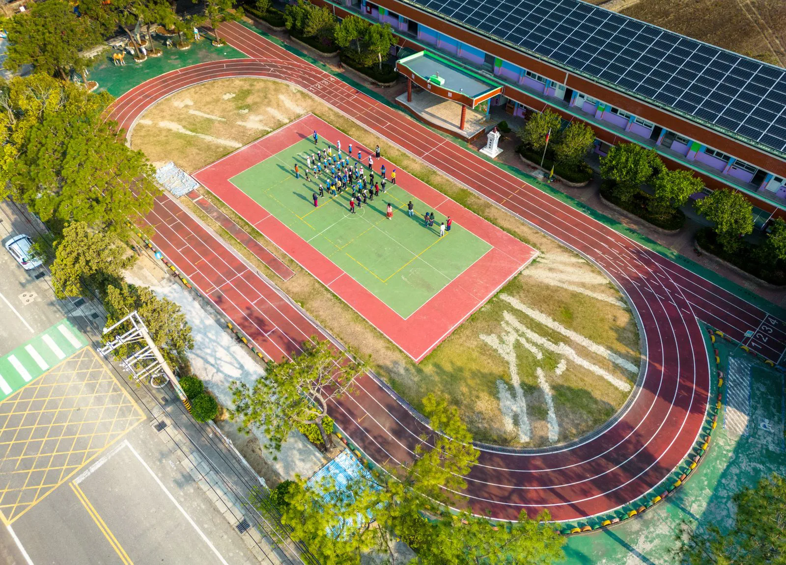 The school sports ground seen from above: a running track around a green field with students assembled