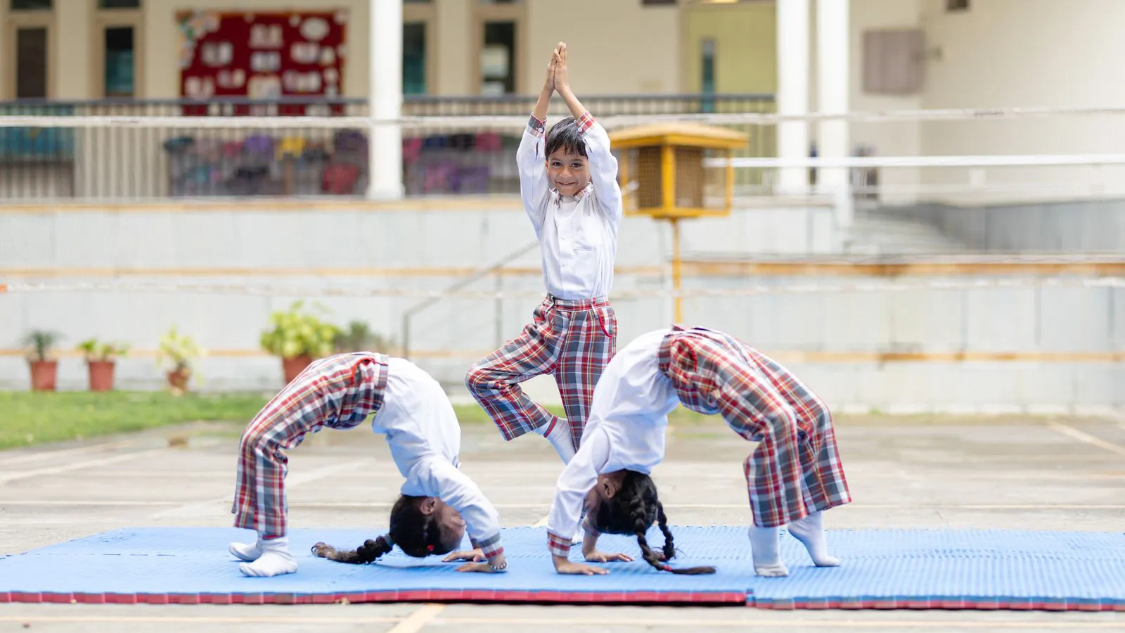 Three children practising yoga poses on a blue mat in the school courtyard
